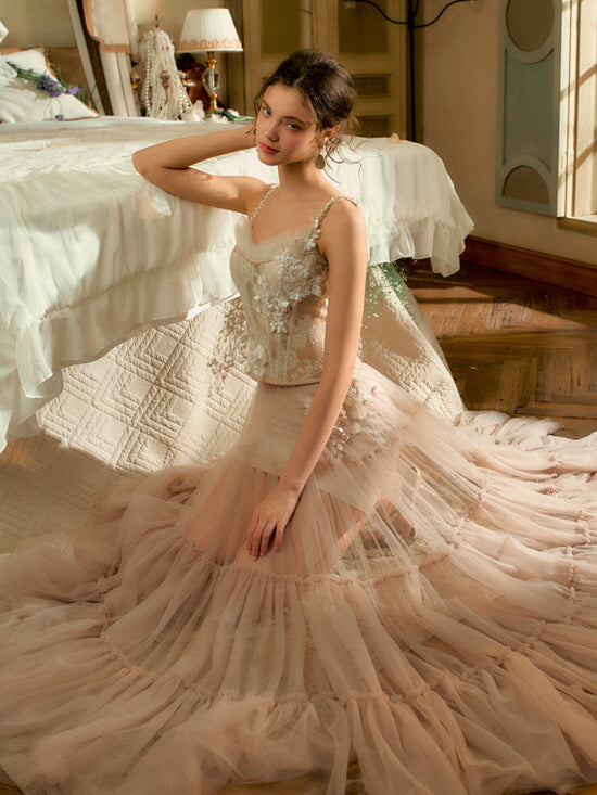 Kneeling beside a romantic bed, the model wears a delicate blush corset and flowy tulle skirt with floral embroidery, evoking a dreamy cottagecore atmosphere.