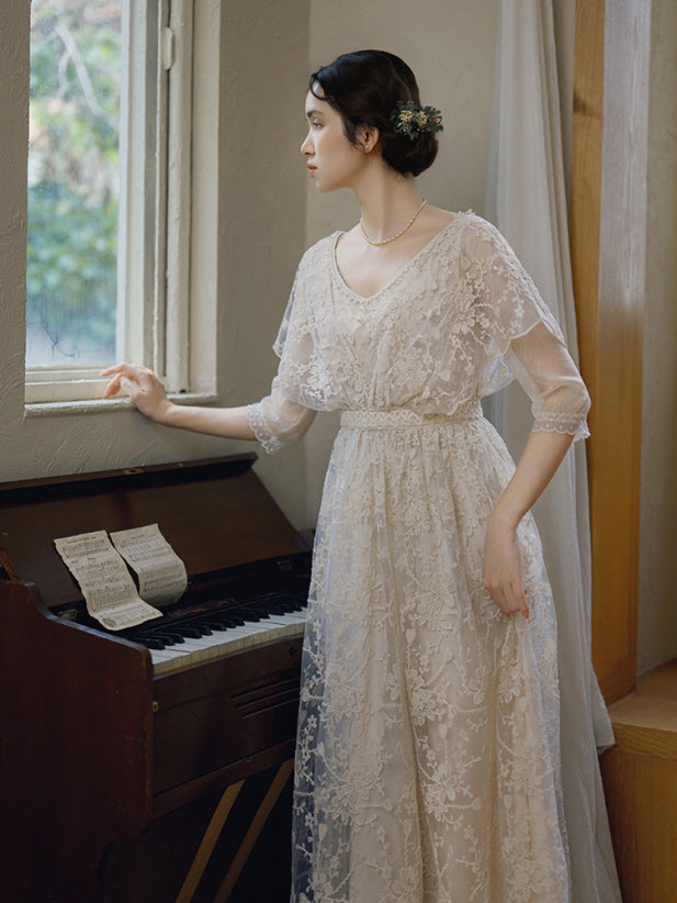 A woman in a delicate embroidered lace dress stands by a window and antique piano, capturing a serene vintage and fairycore moment.