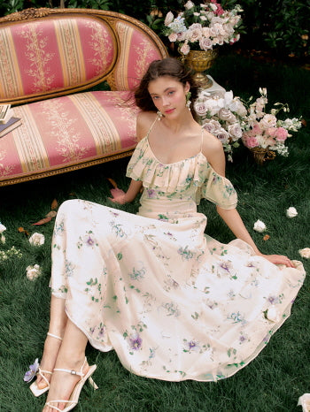 The model sits gracefully on garden grass in a pastel floral dress with cold-shoulder sleeves, embodying a dreamy summer fairycore mood.