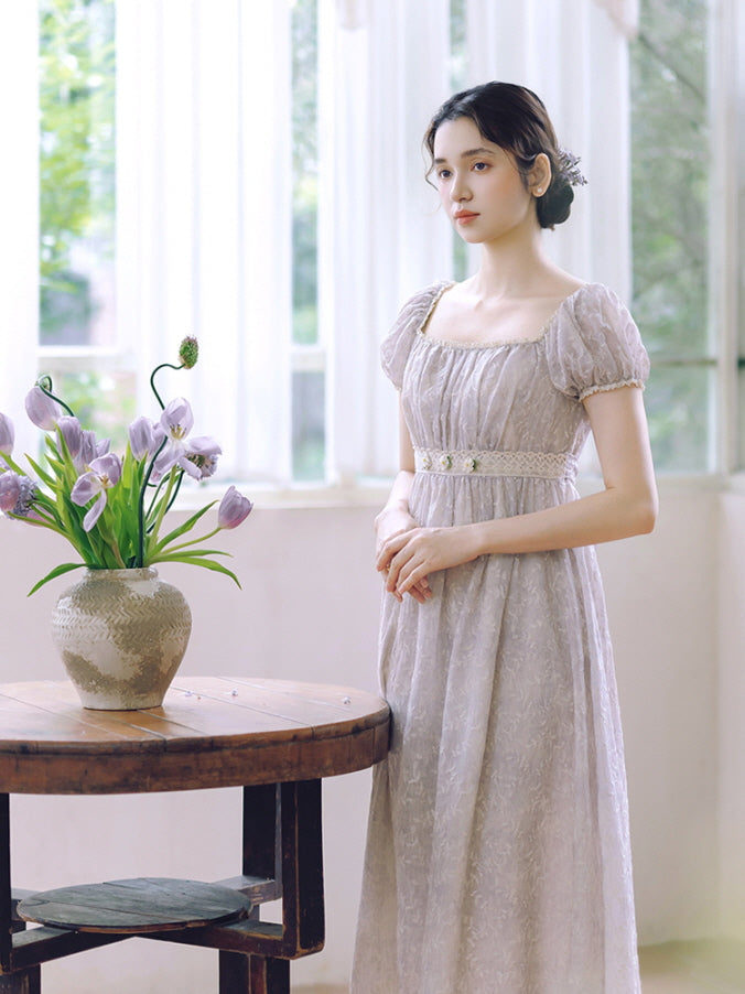A woman stands beside a rustic wooden table in a lavender puff-sleeve dress, channeling a serene vintage cottagecore aesthetic.