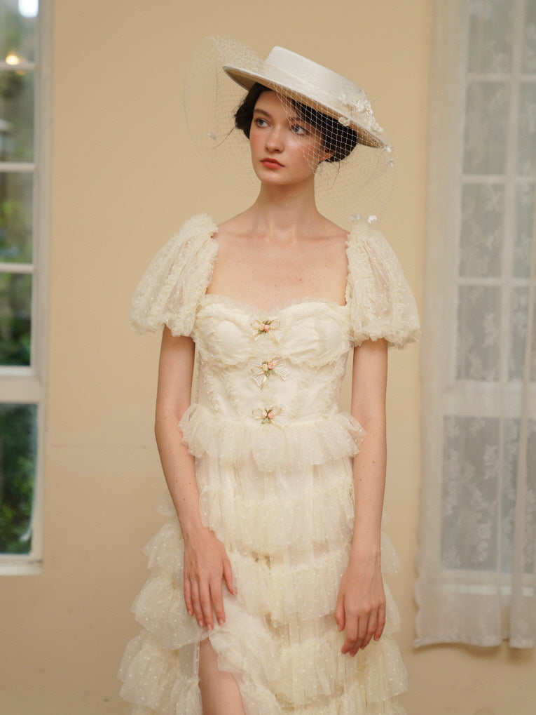 Model in a tiered ivory dress and veiled hat stands near a window, radiating timeless princesscore elegance with delicate puff sleeves and soft lace.