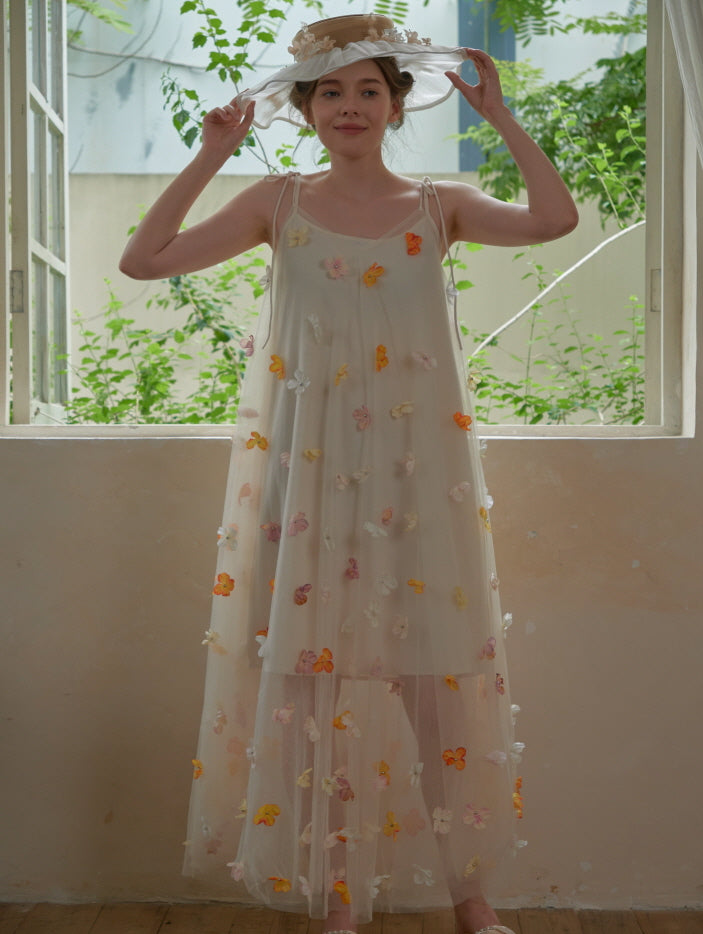 The model adjusts a floral ribbon hat while wearing a dreamy mesh dress covered in colorful petals, glowing in a fairytale-inspired indoor garden.