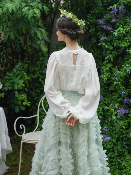 Back view of a woman in a romantic puff-sleeve blouse and layered pastel skirt, styled with a floral crown in a lush princesscore setting.