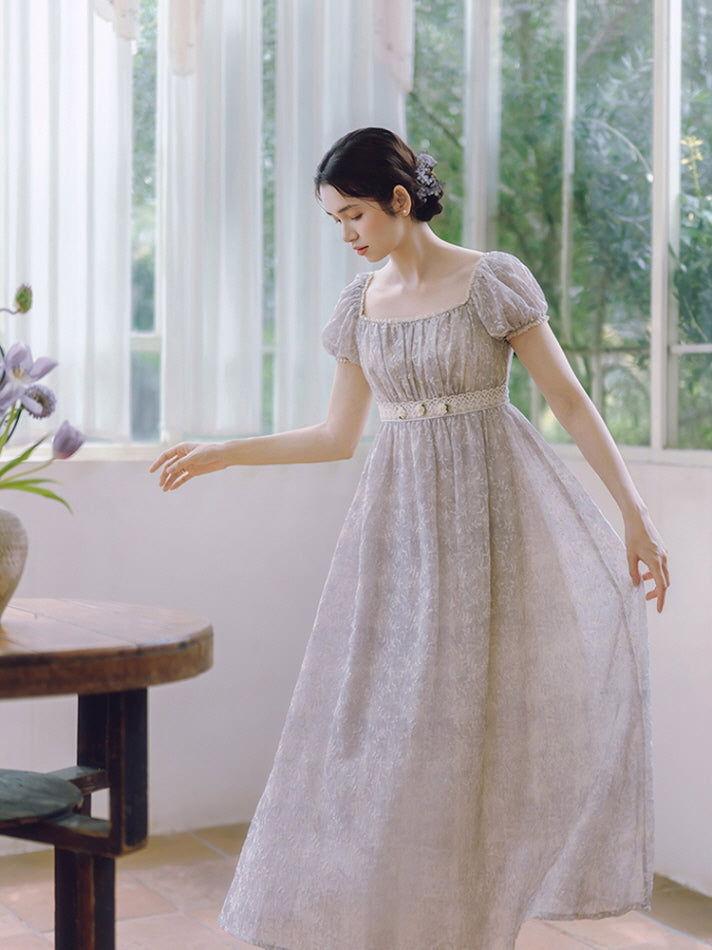In a flowing lavender dress with floral lace detail, a woman gracefully lifts her skirt while surrounded by soft morning light and fairytale charm.