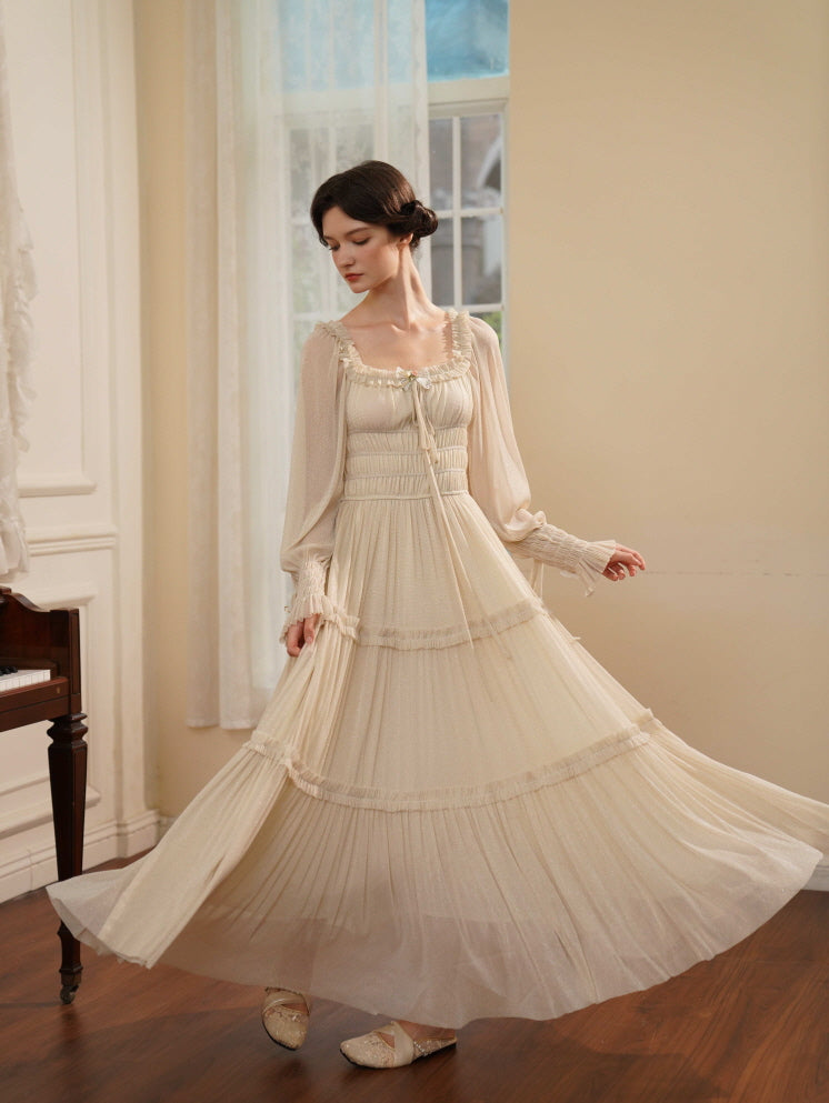 A woman twirls in a flowing cream maxi dress with tiered pleats and long sheer sleeves, radiating elegance in a vintage-inspired outfit.