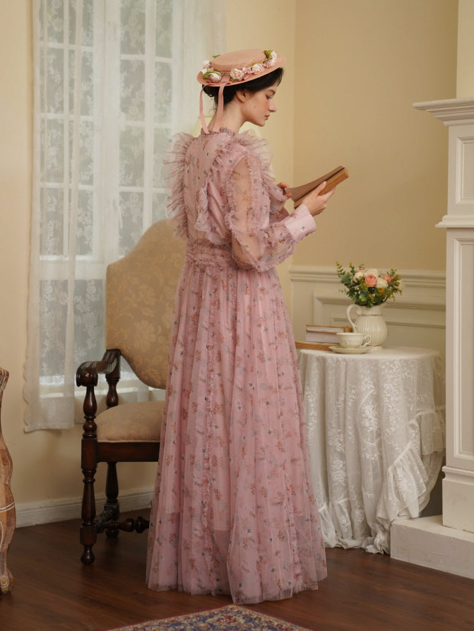 A woman in a pink floral fairycore dress stands beside a lace-covered table, holding a book in a cozy vintage-inspired room.