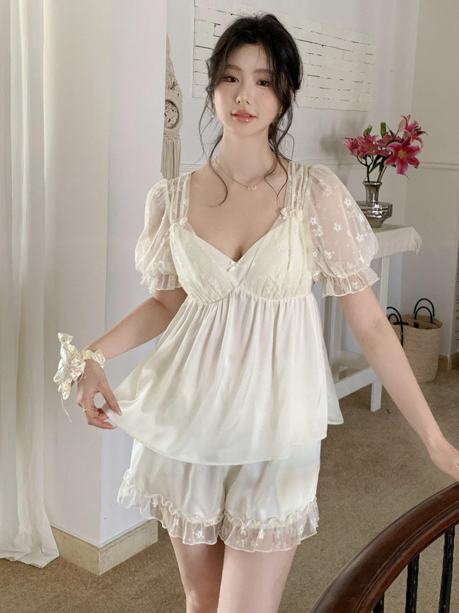 A woman in a white fairycore pajama set with embroidered puff sleeves and ruffled shorts, standing near a staircase in a softly lit vintage room.