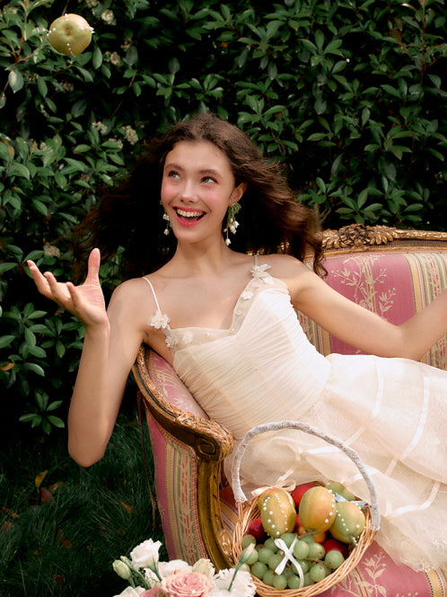 A model in a ruched ivory fairycore dress smiles playfully while tossing fruit on a vintage sofa, surrounded by roses and whimsical charm.