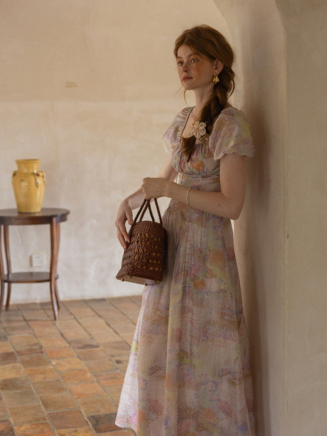 A young woman leans against a wall while holding a woven bag, dressed in a pastel floral dress with puff sleeves, evoking a dreamy, cottagecore look.