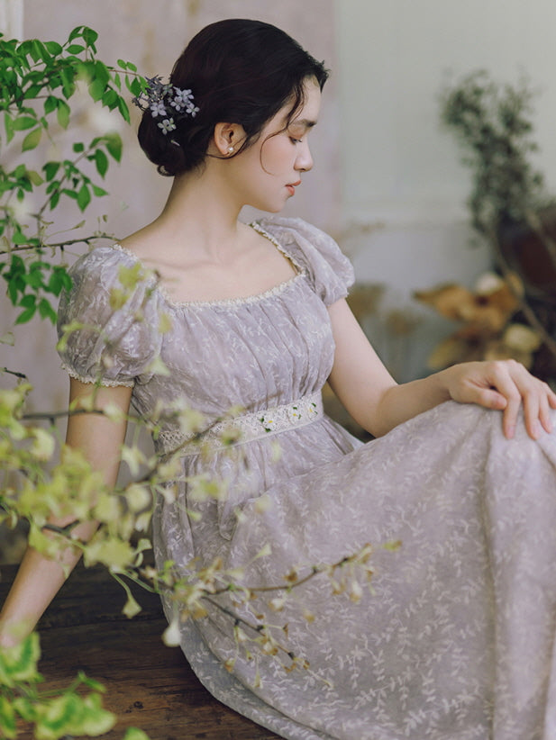 A woman in a soft lavender puff-sleeve dress sits gracefully among greenery, embodying a romantic fairytale and cottagecore atmosphere.