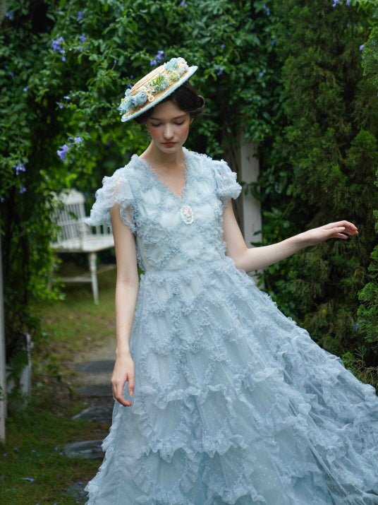 Model in a pale blue ruffled gown gently lifts the hem as she strolls through a blooming garden, evoking soft fairytale romance and timeless charm.