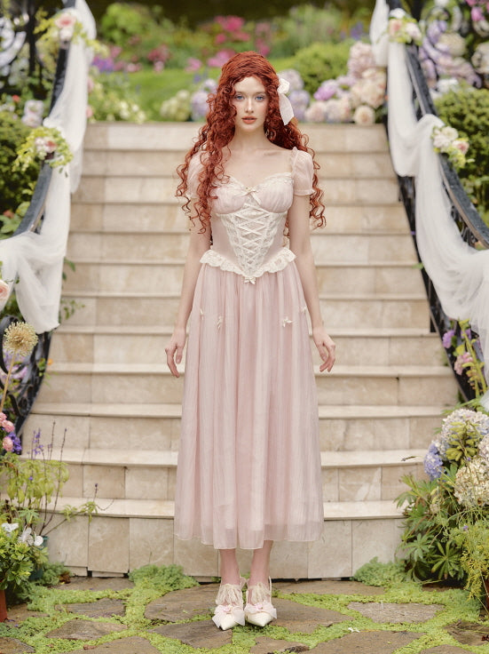 A model with long red curls stands gracefully in a pastel pink corset-style fairytale dress with lace trim and ribbon bows, embodying a princesscore aesthetic.