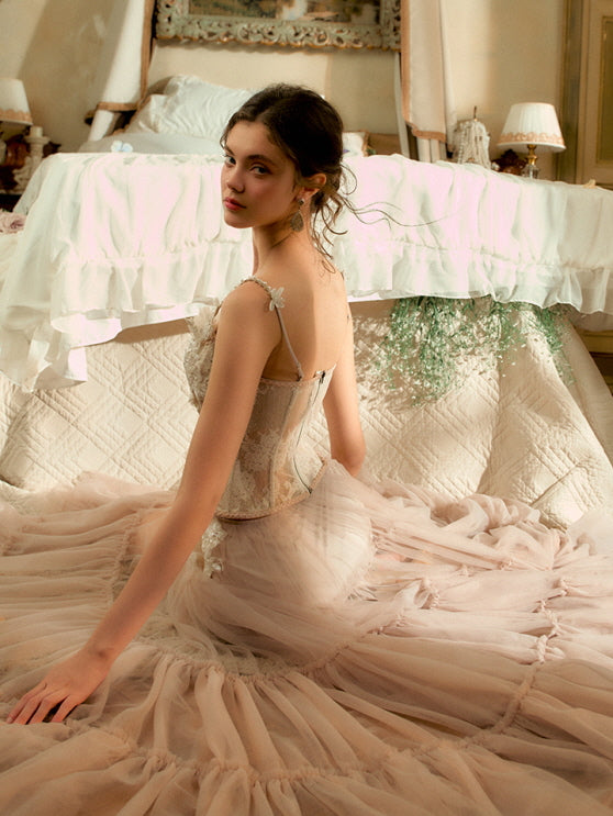 The model sits with her back turned, wearing a sheer blush tulle gown with floral lace corset, surrounded by soft light and romantic vintage decor.