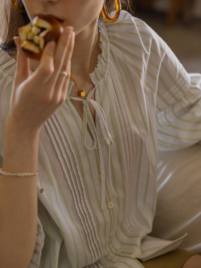 Close-up of a young woman holding a pastry, wearing a light green and white striped blouse with pleated detailing and a tie neckline. The soft, vintage-inspired look exudes a cozy, fairytale charm.