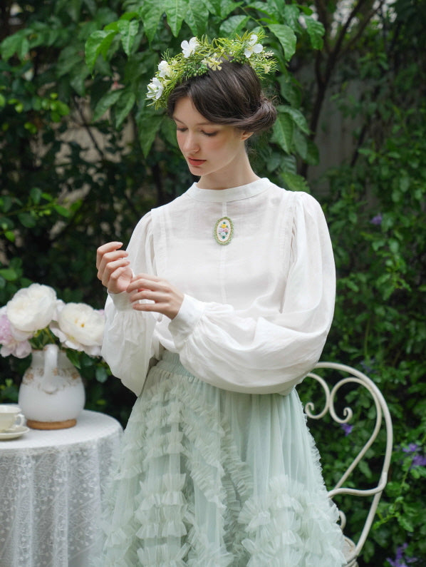 The model wears a white blouse with floral brooch and a mint ruffle skirt, styled with a flower crown in a peaceful garden for a fairycore vibe.