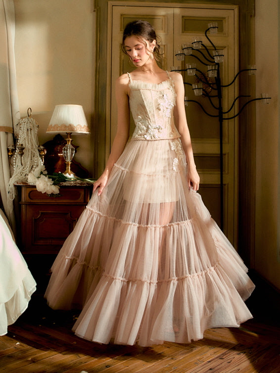 Standing gracefully in a softly lit room, the model gazes down in a layered blush tulle gown with floral embellishments, radiating princesscore elegance.