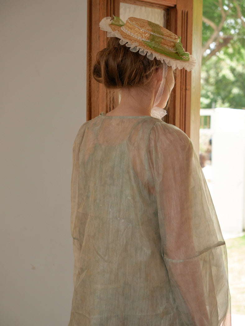 Back view of a woman in a sheer overlay dress and ribbon-trimmed bonnet, standing at the door in a tranquil vintage cottage setting.