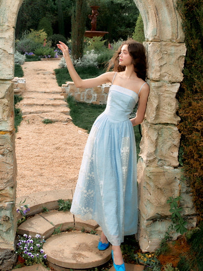 A model lifts her arm gracefully in a pastel blue dress with floral embroidery, framed by a stone arch in a fairytale garden setting.
