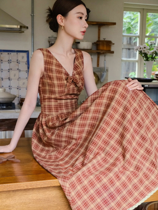 A woman sits in a warm-toned kitchen wearing a red plaid cottagecore dress with a bow detail, capturing a cozy and vintage-inspired mood.