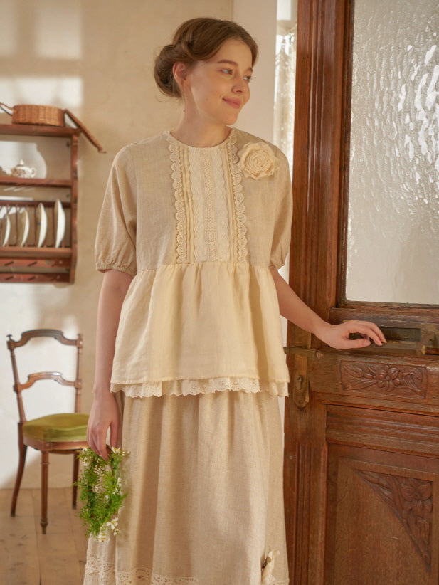 A woman in a beige lace-trimmed top and skirt stands by a carved wooden door, holding a wreath in a soft-lit vintage interior.