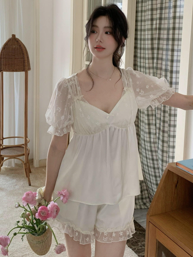 Model holding a vase of pink flowers while wearing a coquette-style ivory sleepwear set with floral mesh sleeves and layered top in a cozy cottage room.