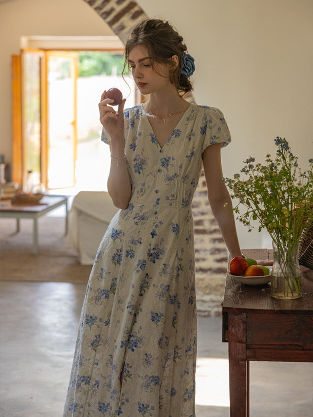 A woman wearing a vintage-inspired cream dress with blue floral embroidery and delicate buttons stands in a rustic room, holding a peach and gazing thoughtfully. The dress exudes a soft cottagecore aesthetic.