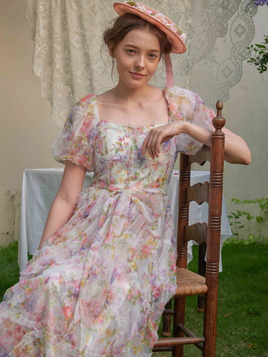 Seated gracefully outdoors, the woman gazes softly while styled in a ruffled fairycore dress with ribbon tie and delicate floral print.