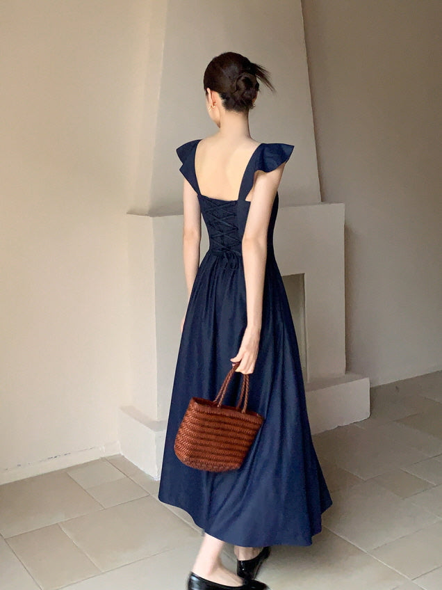 A woman walks gracefully in a princesscore navy dress with a lace-up back, holding a woven bag in a minimalist fairytale-inspired setting.