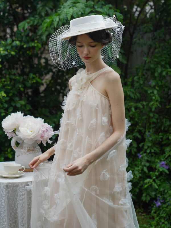 Model in a soft beige halter dress with floral appliqués looks down gently, holding the sheer fabric beside a vintage lace-covered table.
