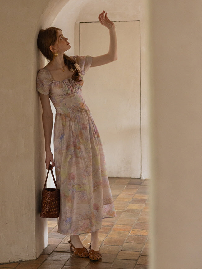 A woman leans against an arched wall while holding a woven bag, dressed in a flowing pastel floral dress with puff sleeves, evoking a dreamy, fairytale vibe.