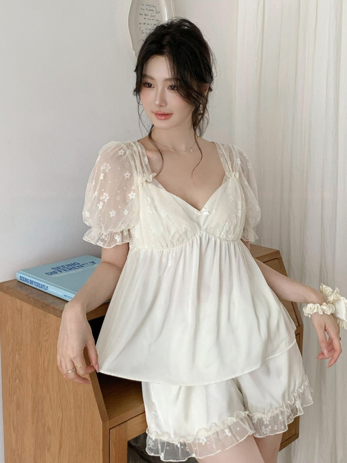 A woman poses beside a wooden desk wearing an ivory coquette pajama set with daisy-embroidered sheer sleeves and ruffle-trim shorts in soft light.