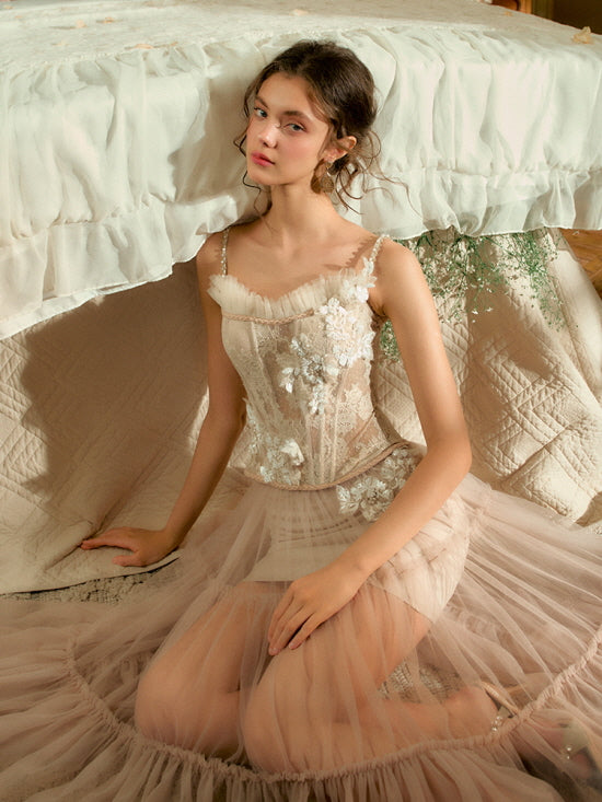 Kneeling beside a vintage bed, the model wears a blush floral corset and layered tulle skirt, capturing a delicate and fairytale-like moment.