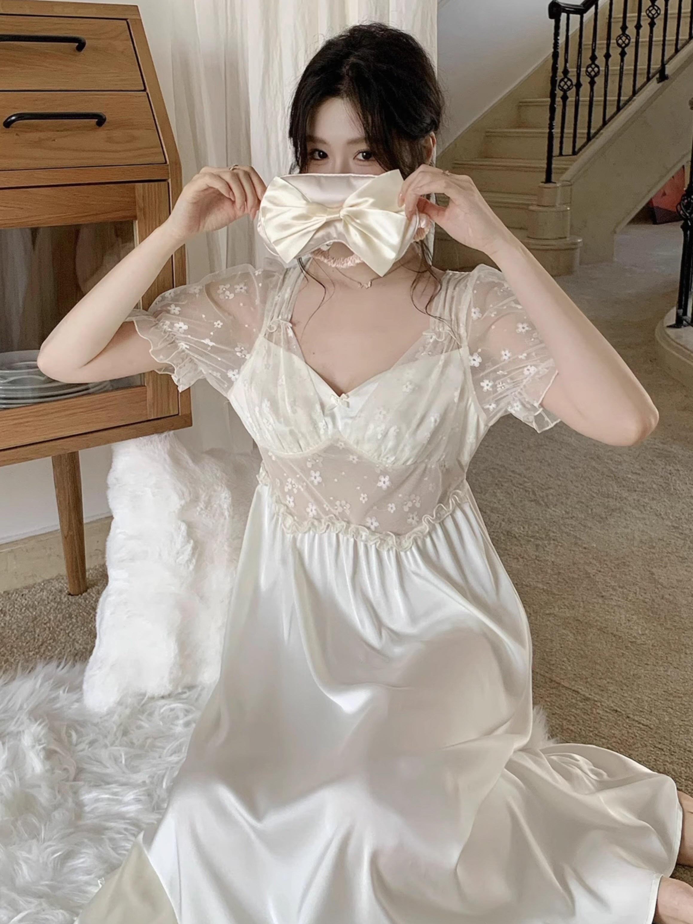 Woman seated on a fluffy rug in an cute  princesscore nightgown, holding a satin bow eye mask with delicate floral embroidery on the bodice.