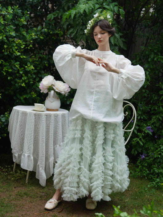 A young woman stands with hands poised in a flowy white blouse and textured mint skirt, evoking a serene fairycore garden aesthetic.