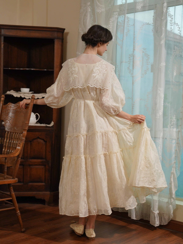 Back view of a woman gently holding the hem of a cream lace dress, standing by sheer curtains in a softly lit vintage-inspired room.