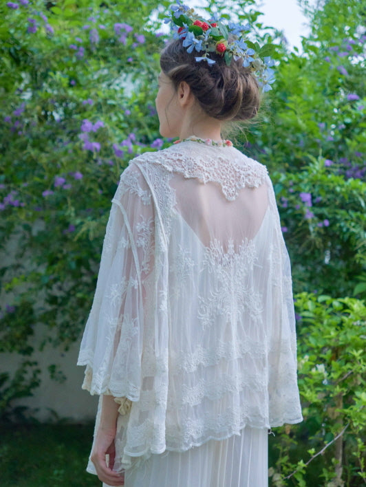 The model shows the back of a delicate embroidered lace robe, styled with a braided updo and flower crown in a cottage garden.