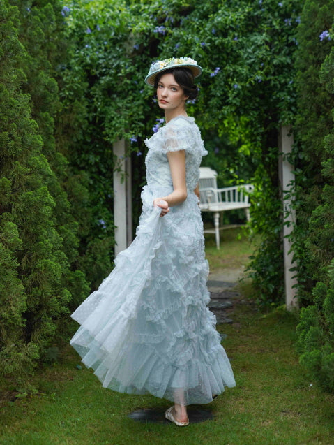 Model turning slightly with a soft gaze, lifting the hem of her ruffled pastel blue dress in a garden path, radiating graceful vintage charm and coquette elegance.
