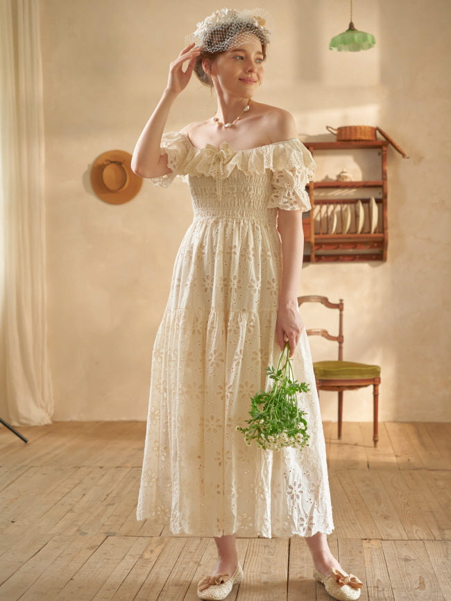 In a vintage-inspired room, a woman in a floral lace dress smiles softly while lifting her hand, holding chamomile flowers in her other hand.
