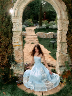 A smiling model sits in a dreamy blue embroidered fairytale dress with spaghetti straps, the full skirt fanned out in a romantic garden setting.