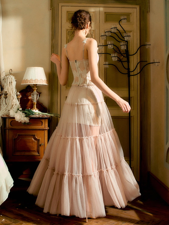 The model walks away from the camera, revealing the delicate back details of a blush corset and layered sheer skirt, evoking timeless fairytale elegance.