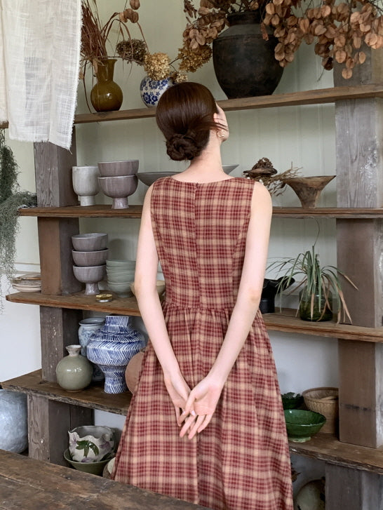 Back view of a woman in a sleeveless gingham dress standing before wooden shelves, evoking a calm and nostalgic cottagecore aesthetic.