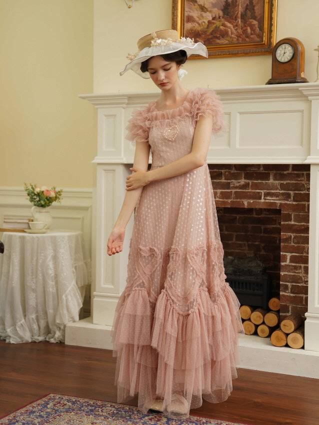 Model poses beside a fireplace wearing a princess-style pink tiered dress with heart accents, evoking a soft and whimsical cottagecore vibe. 