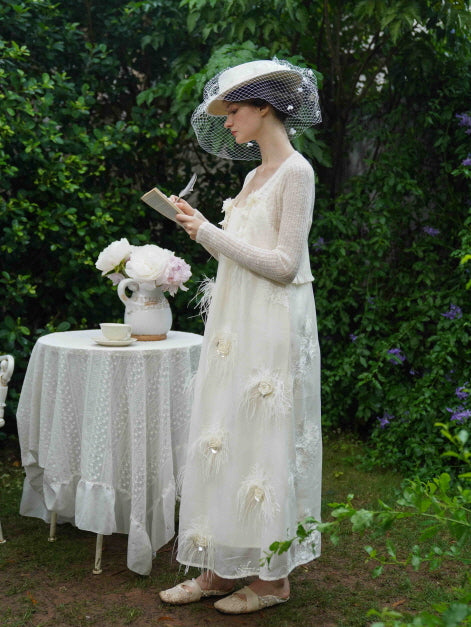 Model writes in a book while wearing a cream embroidered dress with a sheer cardigan, capturing a whimsical cottagecore scene outdoors.