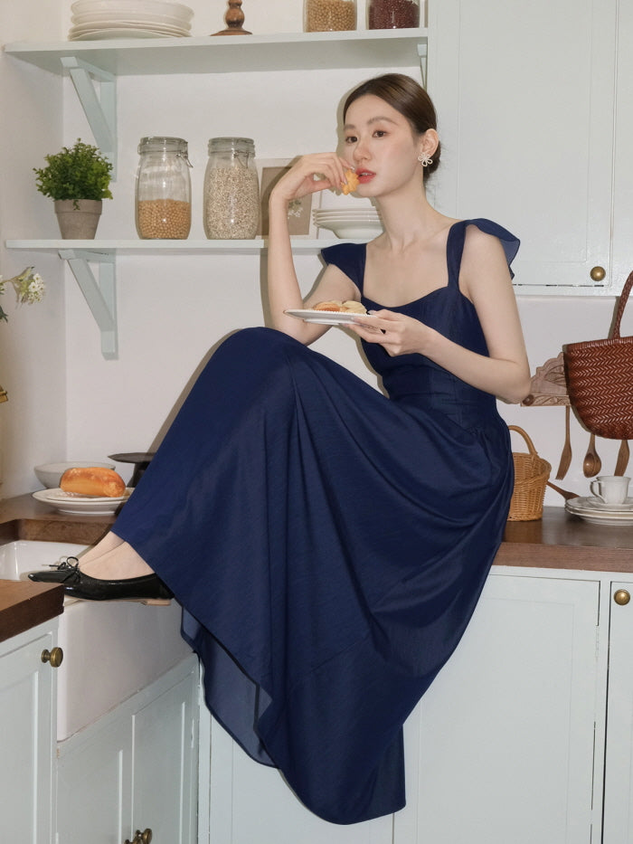 Seated on a cozy kitchen counter, a woman enjoys a pastry in a navy cottagecore dress, blending vintage charm with relaxed fairytale vibes.