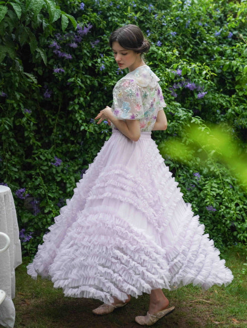 A young woman gracefully twirls in a lavender ruffle skirt and a floral embroidered sheer top, capturing a whimsical princesscore moment in the garden.