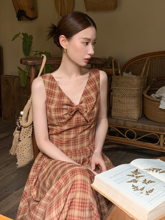 A woman in a red plaid cottagecore dress with a bow detail sits by a wooden desk, gazing softly while holding a botanical book.