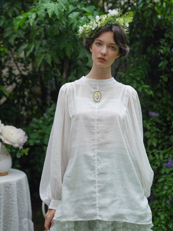 A woman in a white puff-sleeve blouse with a floral brooch gazes softly forward, styled in a vintage garden look with a flower crown.