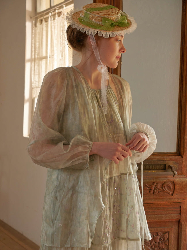 A woman in a sheer puff-sleeve overlay adjusts the waist tie of her pastel green dress, styled with a straw bonnet in soft afternoon light.
