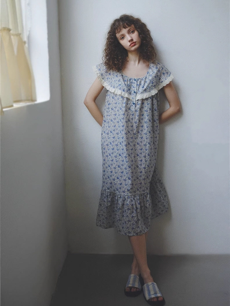 A young woman in a soft blue floral nightgown with lace-trimmed collar leans against a white wall, wearing plaid slippers in a cozy cottagecore look.
