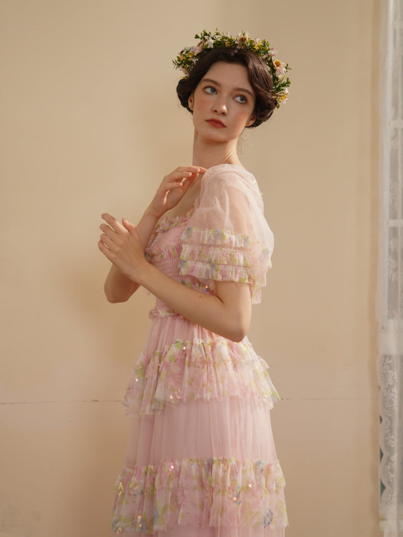 A woman in a pink smocked tulle dress looks upward with a dreamy gaze, wearing a floral crown in a soft vintage fairycore setting.